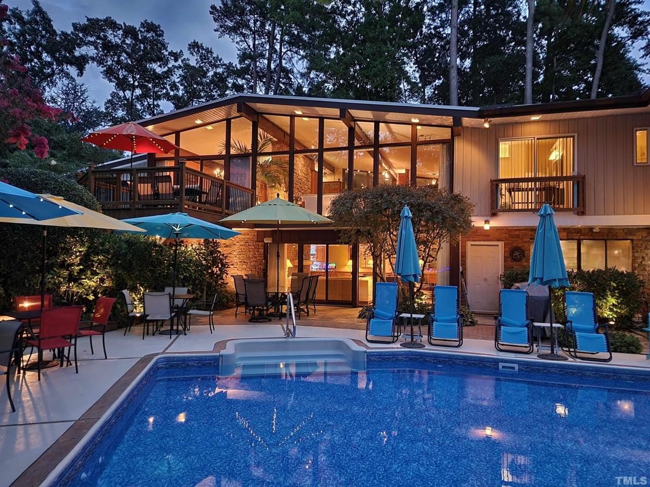 A large home with a crystal blue pool, floor-to-ceiling windows, multiple balconies, and resort-style outdoor furniture, surrounded by trees